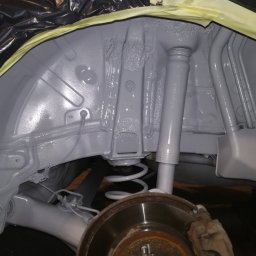 Filtum - Wnętrze nadkola samochodu po zabezpieczeniu antykorozyjnym, widoczna świeża szara powłoka, amortyzator i fragment tarczy hamulcowej, krawędzie zabezpieczone taśmą malarską.