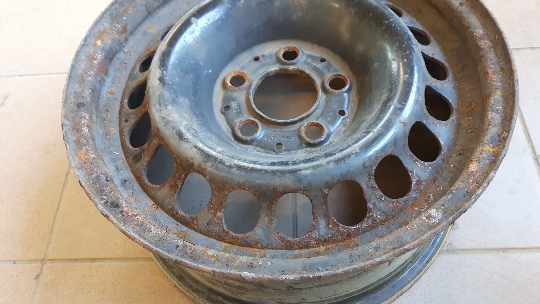 Stara stalowa felga samochodowa z widoczną rdzą na krawędziach i powierzchni, leżąca na jasnych płytkach ceramicznych.