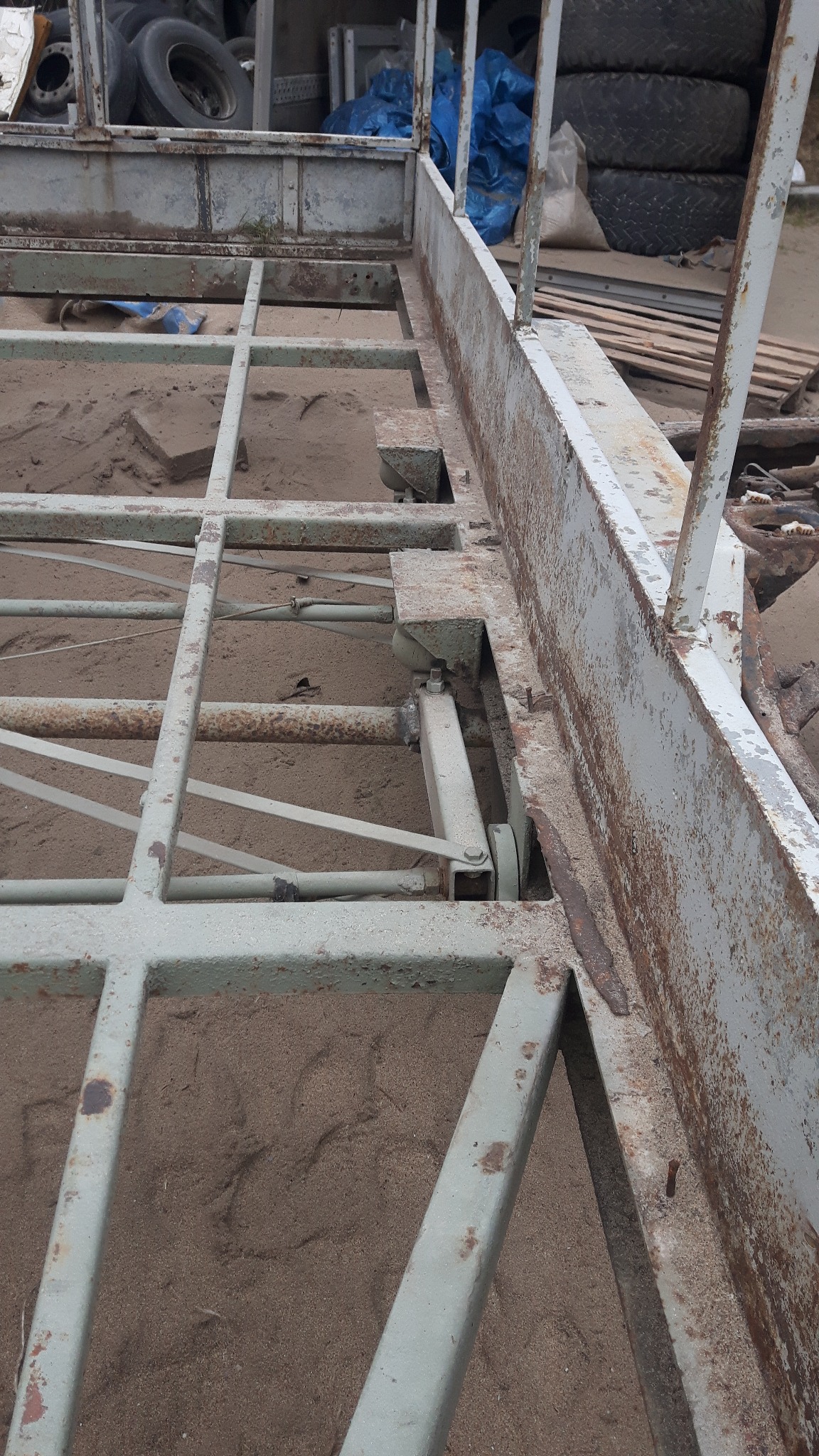 Rama przyczepy pokryta rdzą i piaskiem, widoczne ślady korozji i spawania, opony w tle.