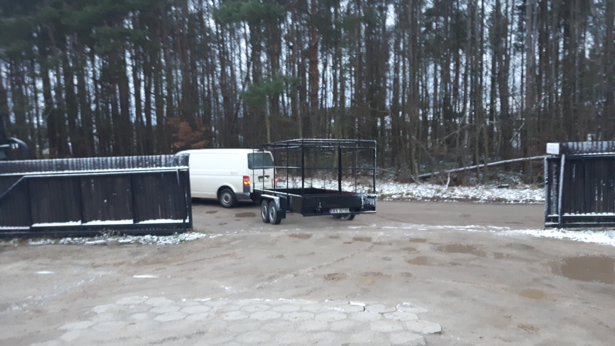 Czarna, metalowa przyczepka samochodowa z klatką, zaparkowana na błotnistym placu w zimowy dzień, widoczny biały samochód dostawczy i las w tle.