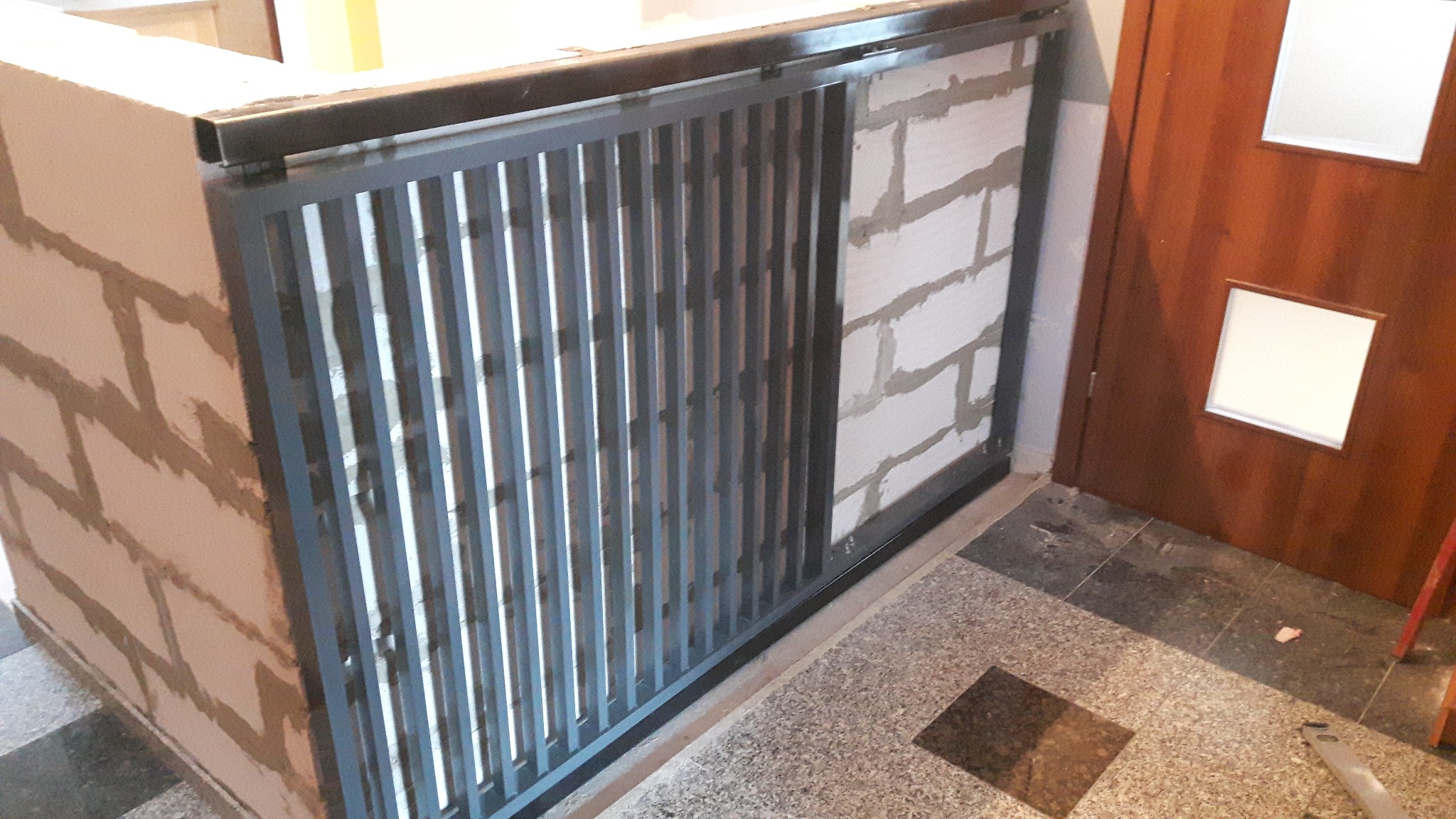 Szara metalowa balustrada z pionowymi szczebelkami, zamontowana na murze z białych bloczków, widok z dołu na fragment klatki schodowej z terazzo i brązowe drzwi z białymi szybkami.