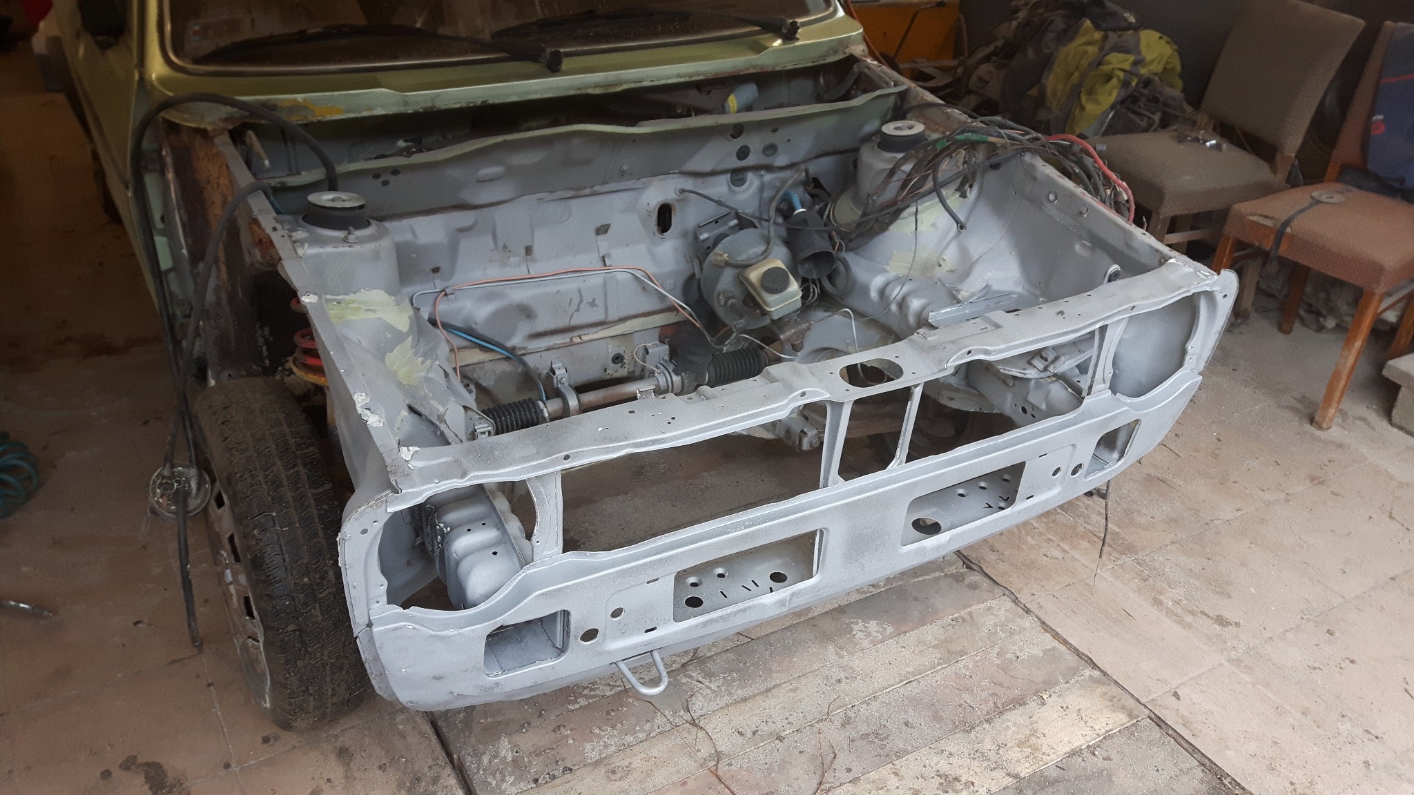 Przód samochodu w trakcie renowacji, z widocznym odnowionym nadwoziem w kolorze szarym, odsłoniętym silnikiem i elementami zawieszenia, na tle warsztatu z drewnianą podłogą i krzesłami.