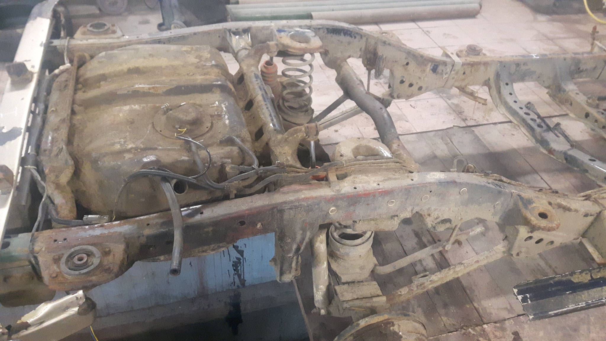 Zardzewiała rama samochodu z widocznym zbiornikiem paliwa i elementami zawieszenia, ukazująca zaawansowany stan korozji na tle drewnianej podłogi warsztatu.