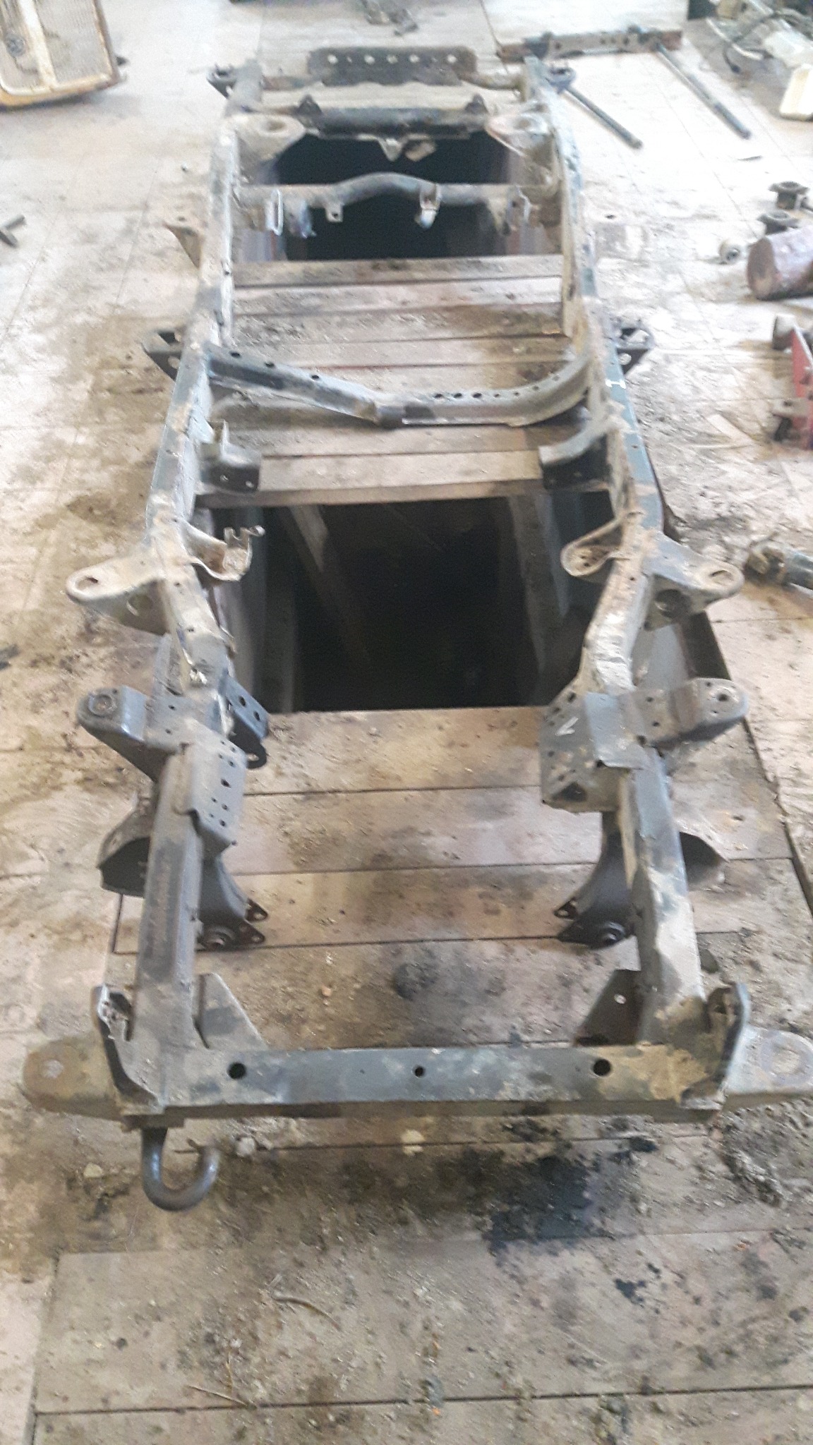 Metalowa rama pojazdu po demontażu, widoczne ślady korozji i zabrudzenia, ułożona na drewnianej podłodze warsztatu.