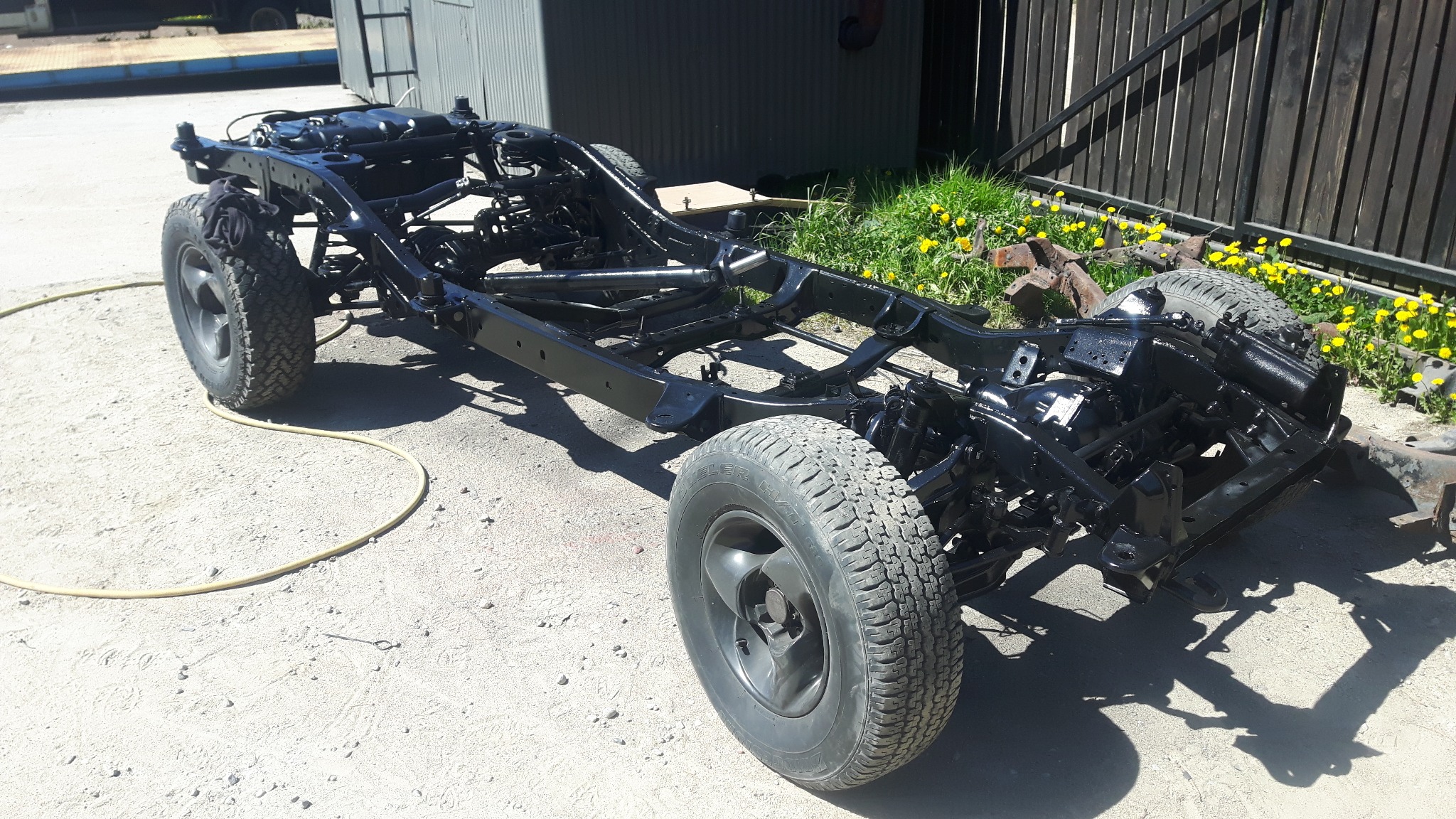 Podwozie samochodu po renowacji, świeżo pomalowane na czarno, z widocznymi oponami, na tle betonowego placu i ogrodzenia.