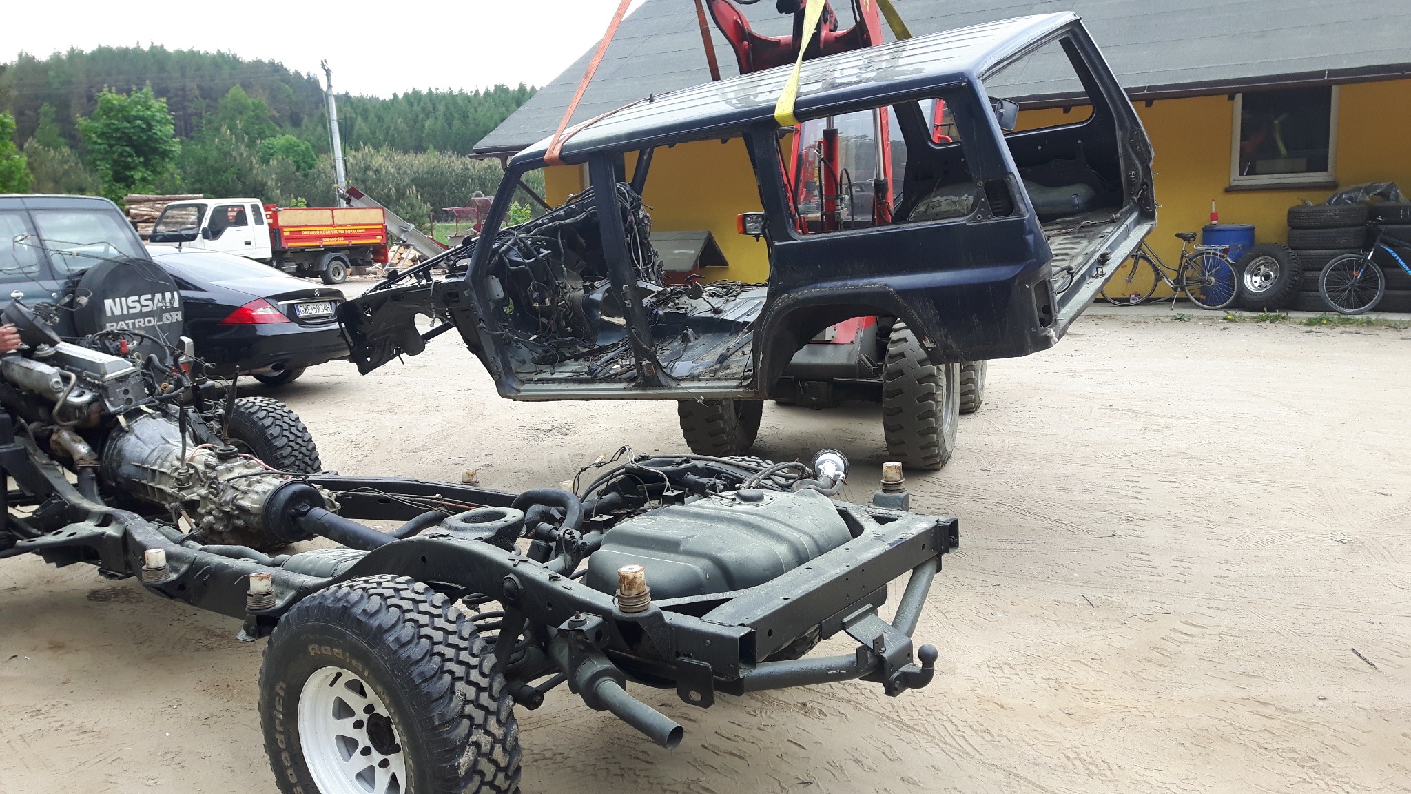 Demontaż karoserii samochodu terenowego Nissan Patrol zawieszonej na pasach dźwigu, widoczna rama z silnikiem i skrzynią biegów, w tle zaparkowane samochody i sterta opon.