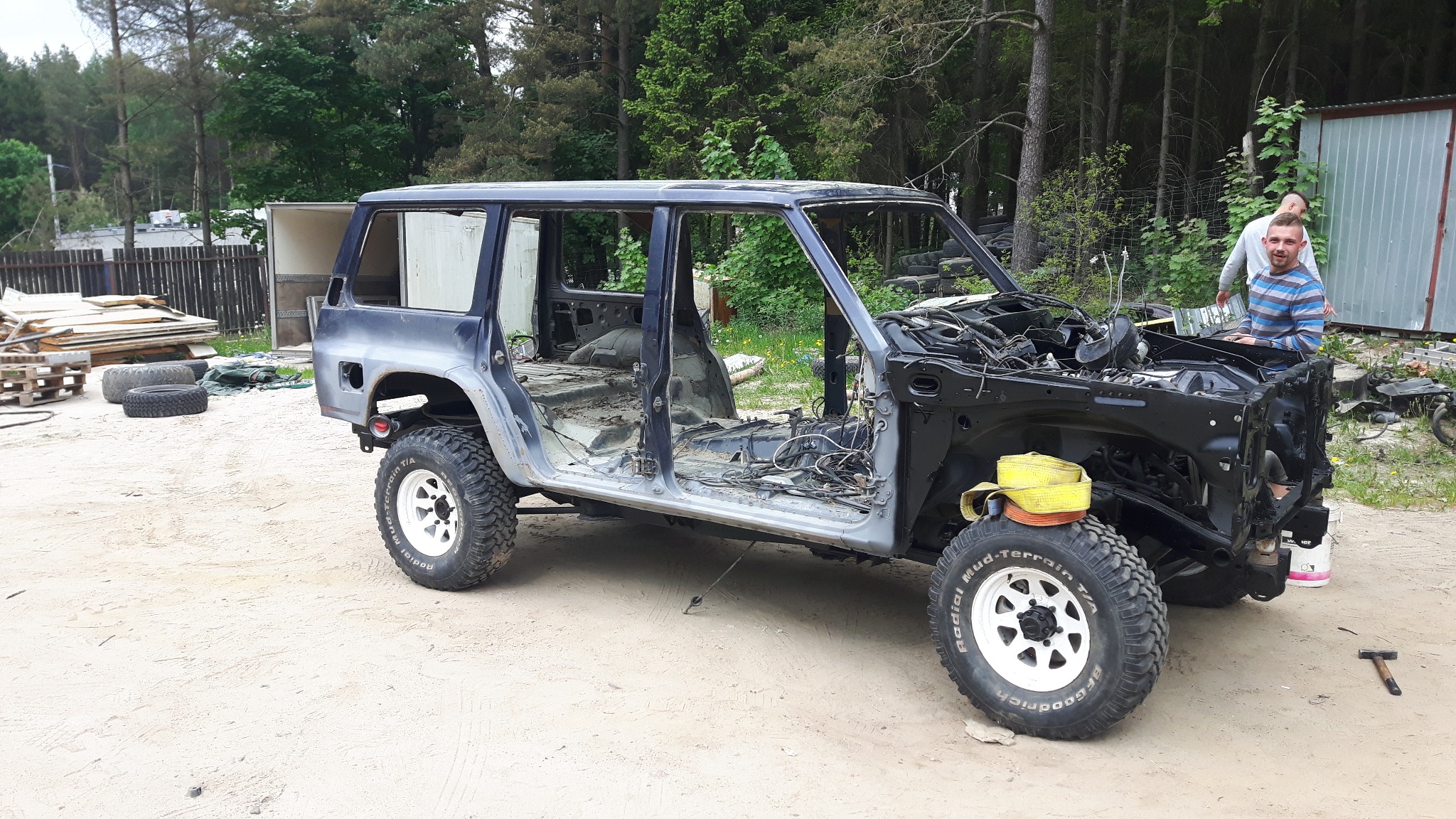 Demontaż karoserii samochodu terenowego z widocznymi przewodami i elementami silnika, dwóch mężczyzn w tle przy pojeździe, teren piaszczysty.