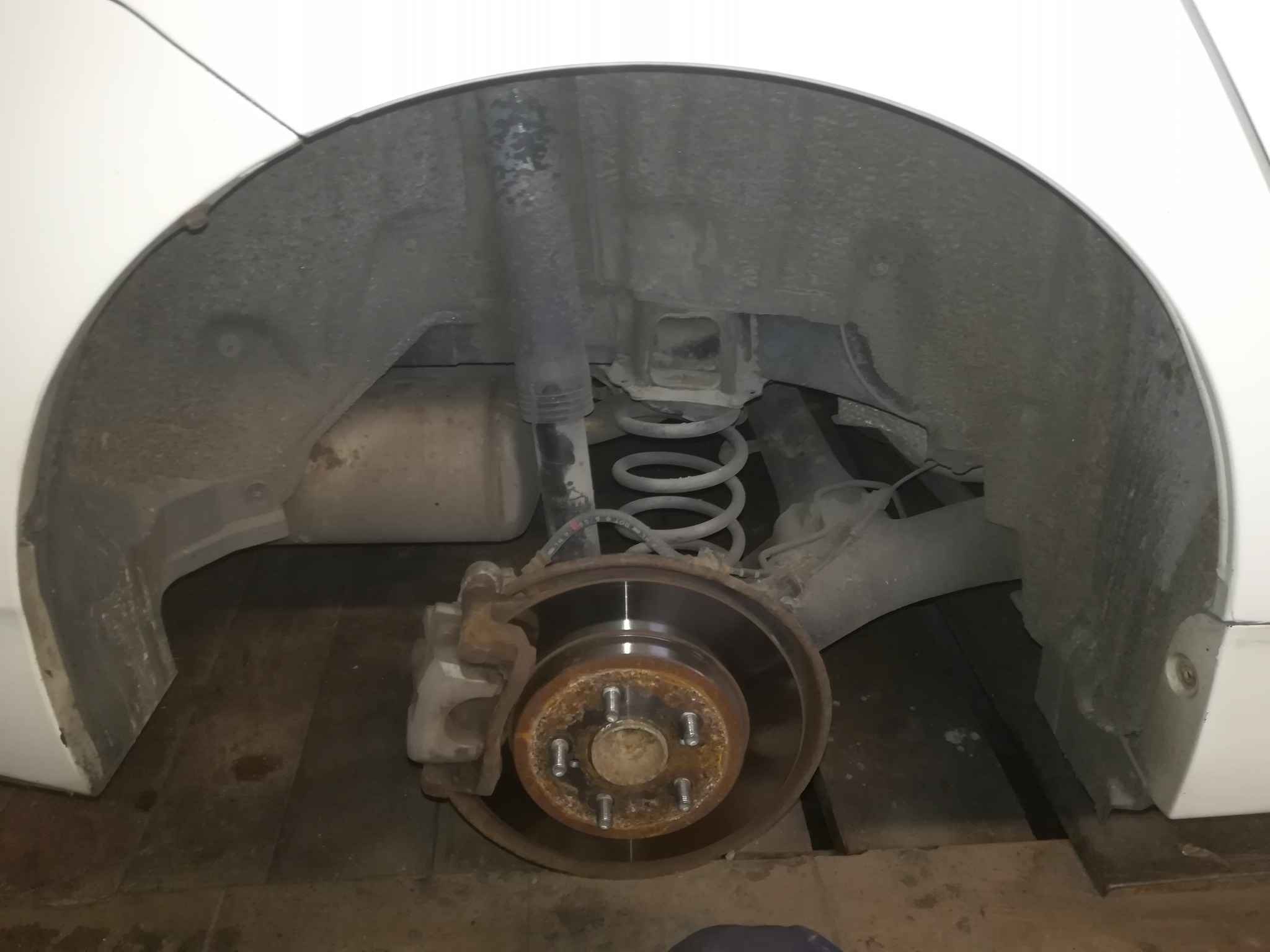 Widok na tylne zawieszenie samochodu z widoczną tarczą hamulcową, sprężyną i amortyzatorem, zardzewiałe śruby mocujące koło.