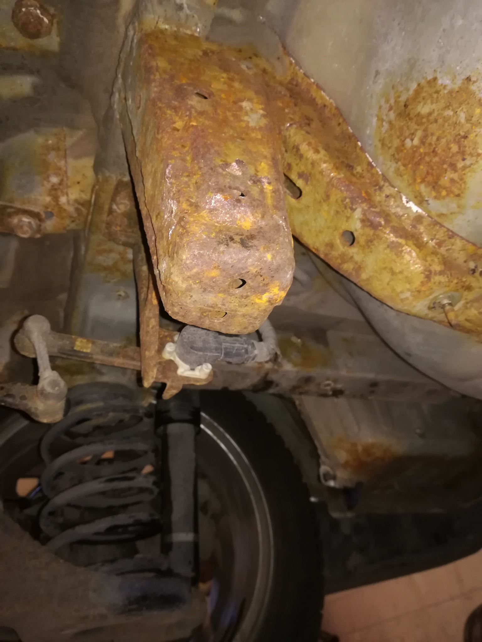 Silnie skorodowany element podwozia samochodu, widoczne ślady rdzy na metalowych częściach, sprężyna zawieszenia i opona w tle.