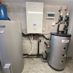 Pompy Ciepła Brodnica - Zestaw urządzeń grzewczych: zbiornik Ferroli z niebieskim naczyniem wzbiorczym, biała jednostka Panasonic z wyświetlaczem, zbiornik WHS z izolacją, połączone rurami w pomieszczeniu z szarą glazurą.