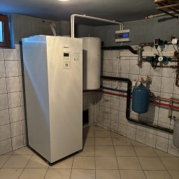 Pompy Ciepła Brodnica - Biała pompa ciepła Panasonic Aquarea w kotłowni z ceramiczną podłogą i ścianami w płytkach, widoczne rury instalacji hydraulicznej i elektryczna skrzynka rozdzielcza.