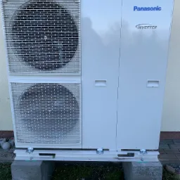 Biała pompa ciepła Panasonic Inverter, zamontowana na betonowych blokach przy ścianie budynku, widoczne dwie kratki wentylacyjne.