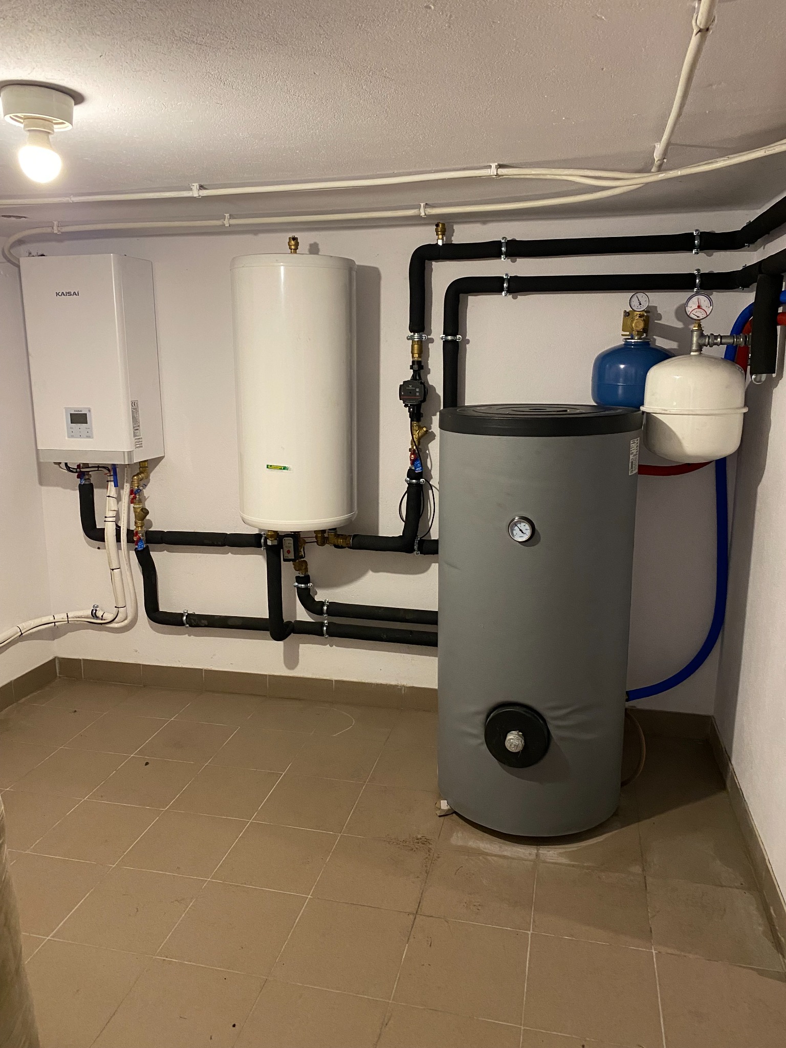Zestaw urządzeń grzewczych w kotłowni: wiszący piec Kaisai z wyświetlaczem, biały zasobnik ciepłej wody, szary zbiornik buforowy z termometrem, połączone czarnymi rurami w izolacji, niebieskie...