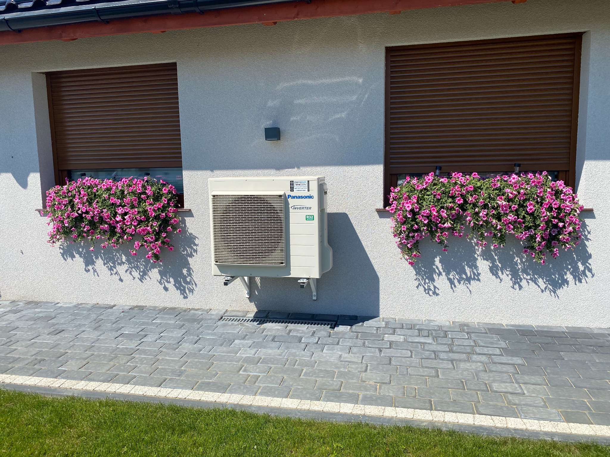 Biała jednostka zewnętrzna klimatyzacji Panasonic Inverter zamontowana na elewacji domu pomiędzy dwoma oknami z brązowymi roletami i skrzynkami z różowymi kwiatami.