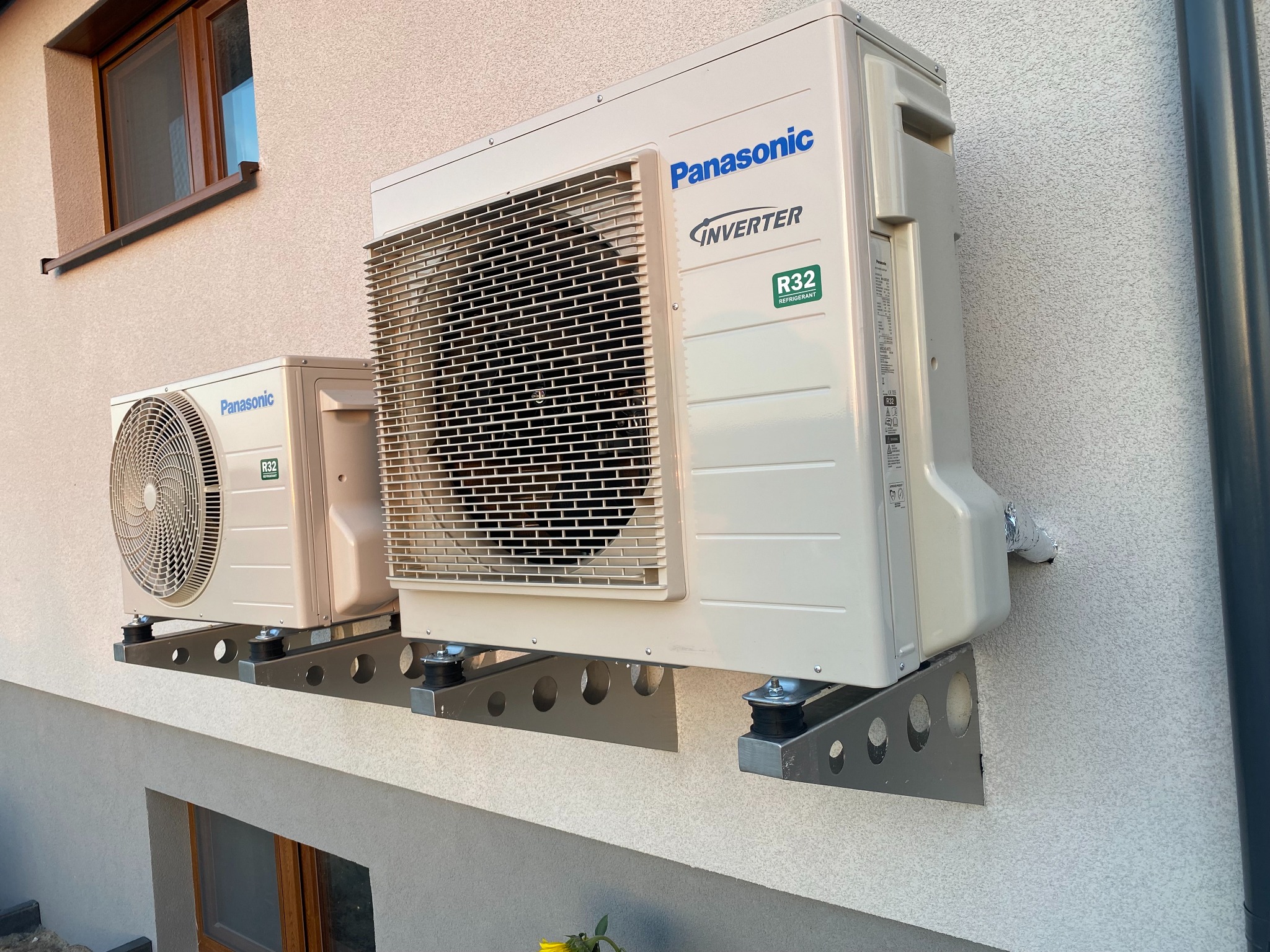 Dwa agregaty klimatyzacji Panasonic Inverter R32 zamontowane na metalowych wspornikach z otworami, przymocowane do elewacji budynku z oknem.