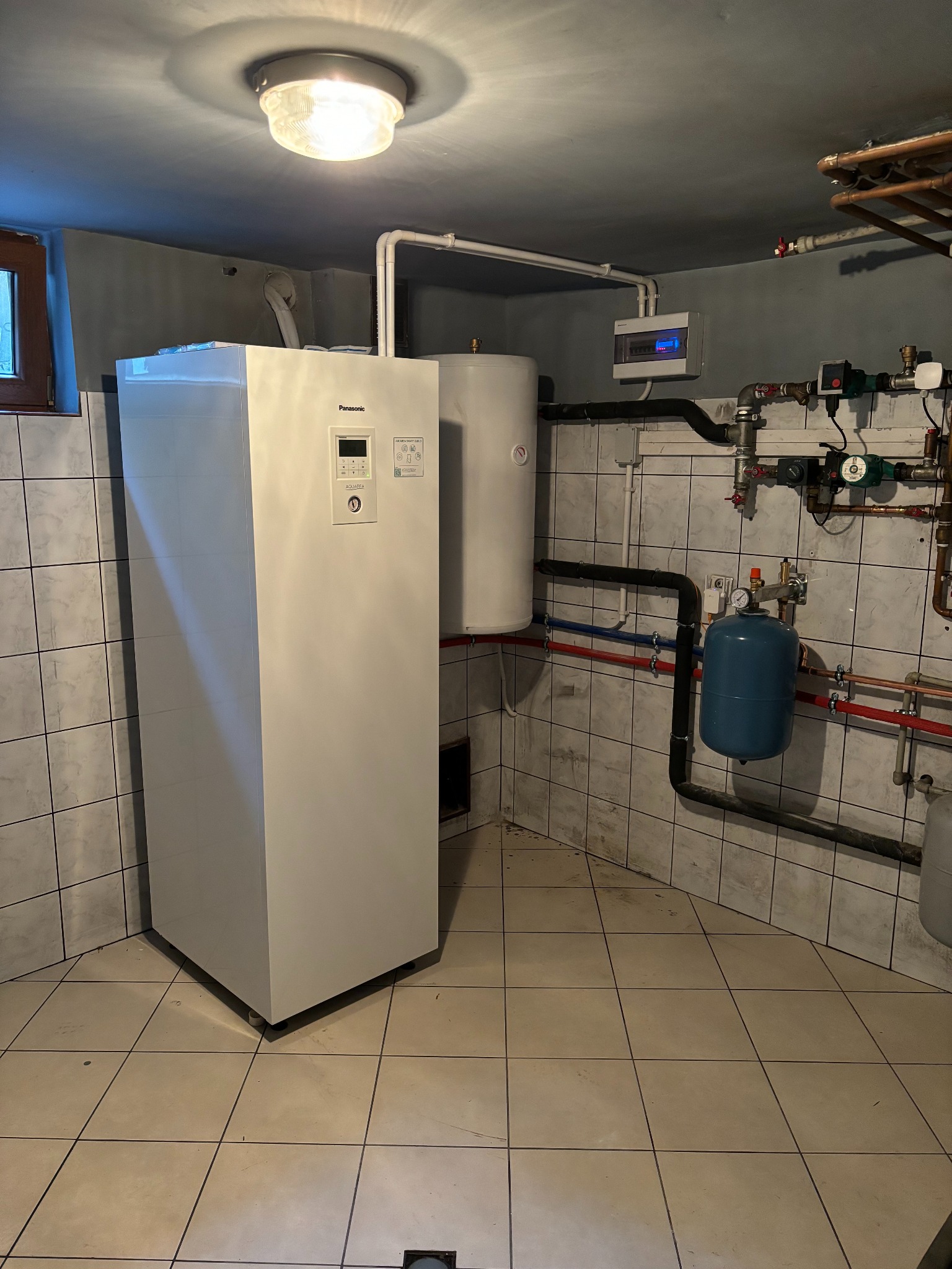 Biała pompa ciepła Panasonic Aquarea w kotłowni z ceramiczną podłogą i ścianami w płytkach, widoczne rury instalacji hydraulicznej i elektryczna skrzynka rozdzielcza.