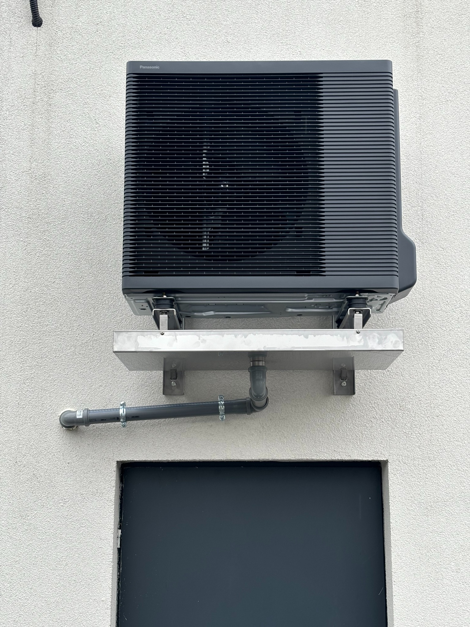 Zamontowana na zewnątrz jednostka klimatyzacji Panasonic na metalowym wsporniku, z widocznym odpływem kondensatu i fragmentem ściany z oknem.