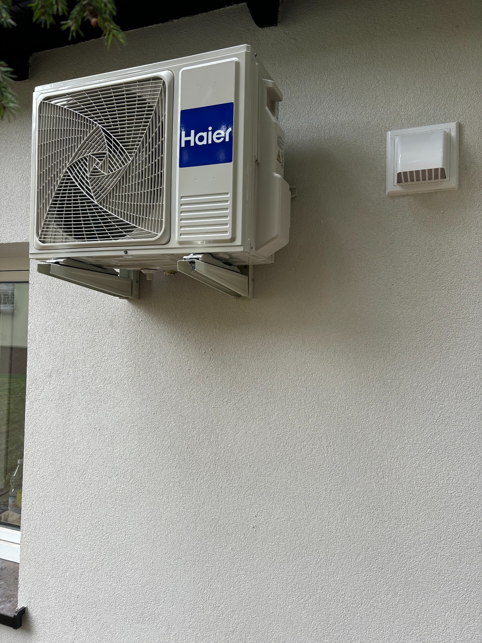 Biała jednostka zewnętrzna klimatyzatora Haier zamontowana na wspornikach na szarej, teksturowanej ścianie budynku, obok kratka wentylacyjna.