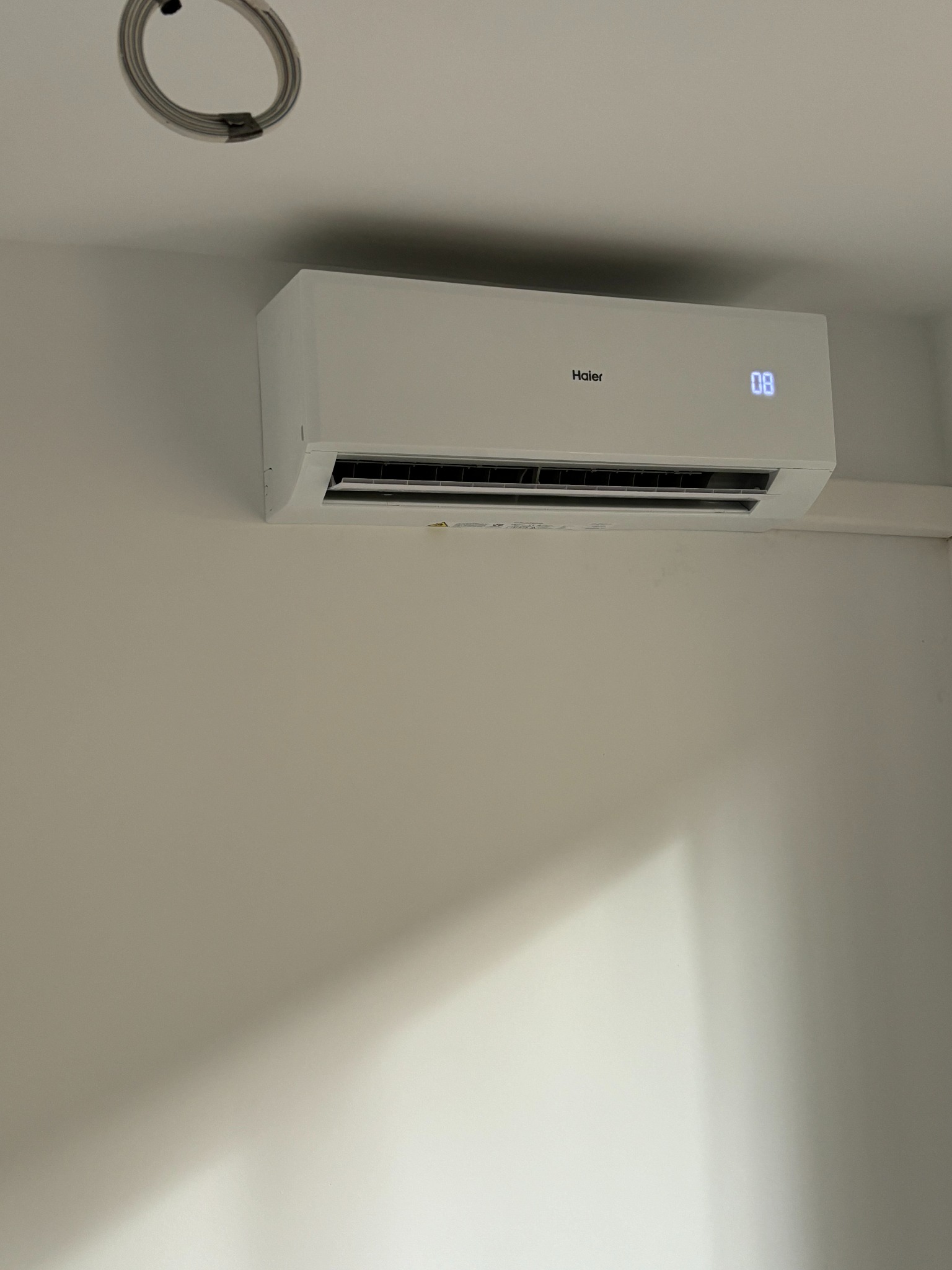 Biała jednostka klimatyzacyjna Haier zamontowana na ścianie w jasnym pomieszczeniu, widoczny wyświetlacz z temperaturą i częściowo odsłonięte przewody elektryczne w suficie.
