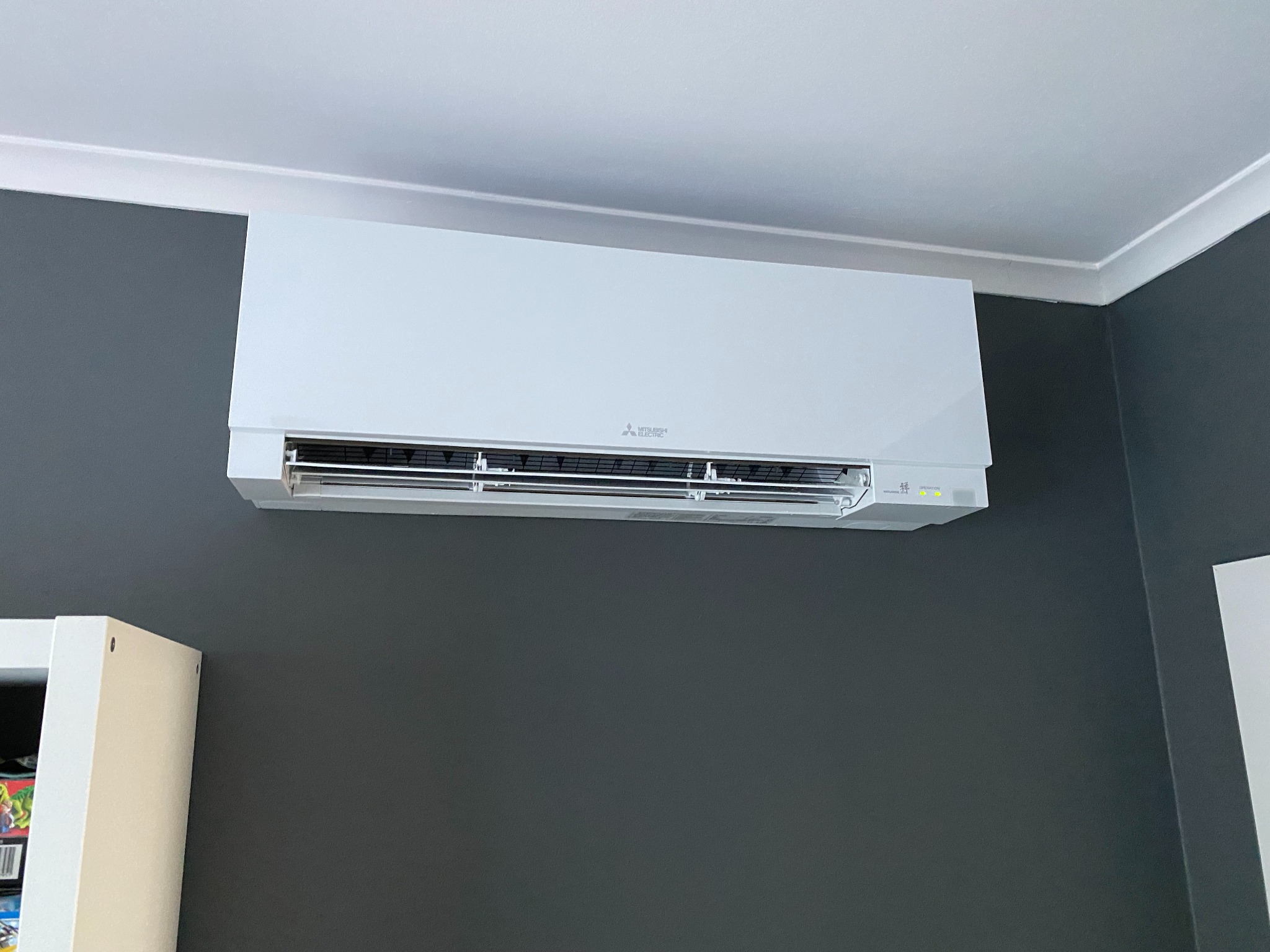 Biała klimatyzacja ścienna marki Mitsubishi Electric zamontowana na szarej ścianie w pomieszczeniu z białym sufitem i listwą przysufitową, widoczny fragment białego regału z lewej strony.