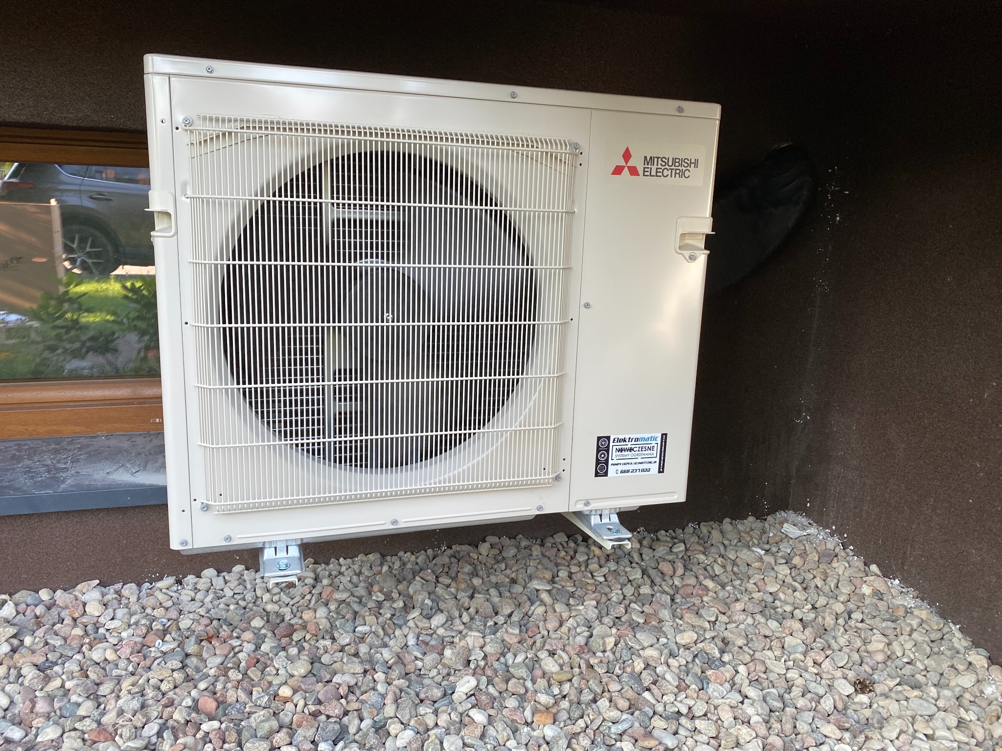 Zewnętrzna jednostka klimatyzacji Mitsubishi Electric zamontowana na elewacji domu, posadowiona na żwirowym podłożu, widoczny fragment okna i odbicie samochodu.