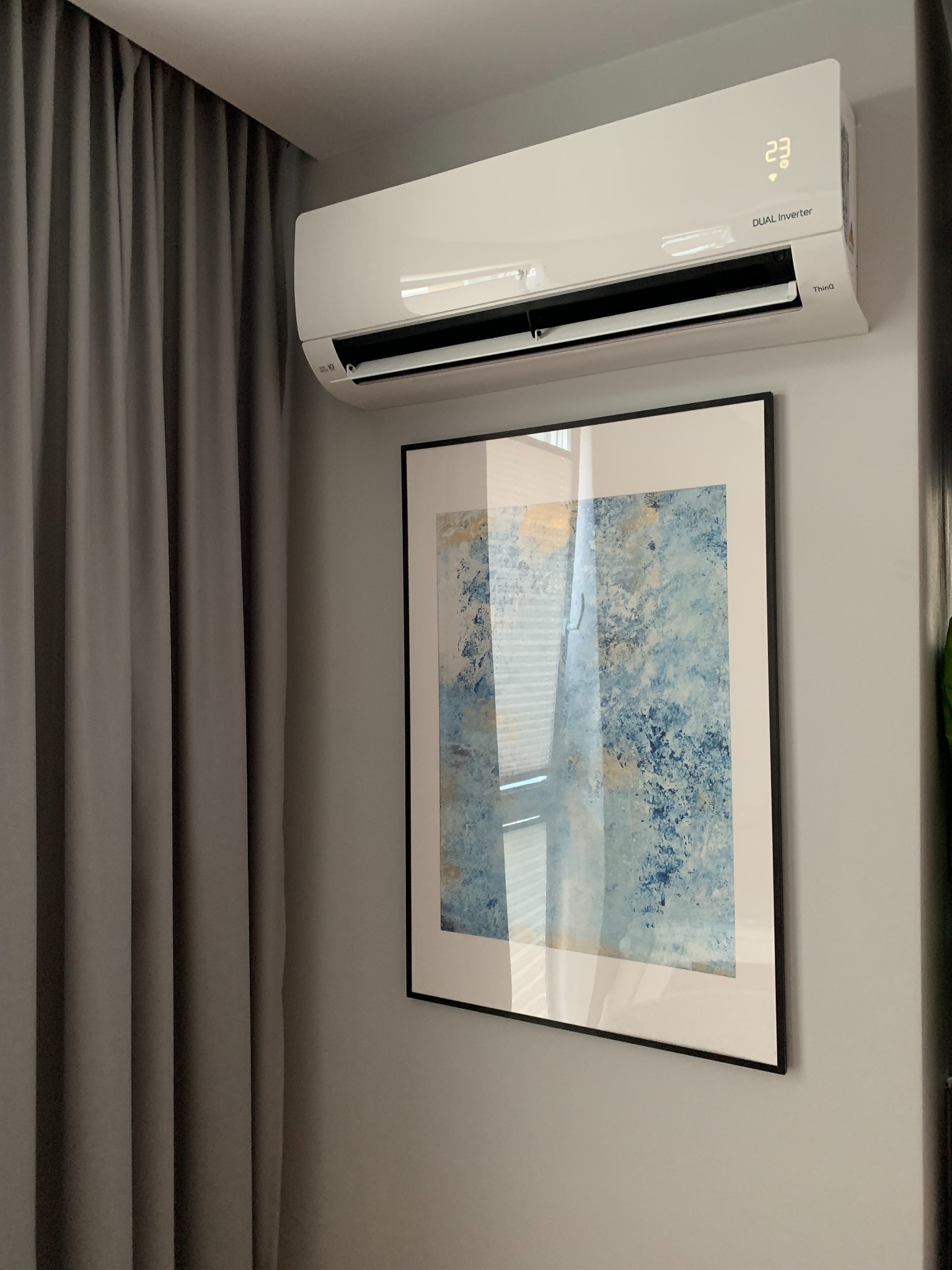 Biały klimatyzator ścienny LG Dual Inverter z wyświetlaczem temperatury 23 stopnie Celsjusza, zamontowany nad obrazem w czarnej ramie na szarej ścianie, obok szarych zasłon.