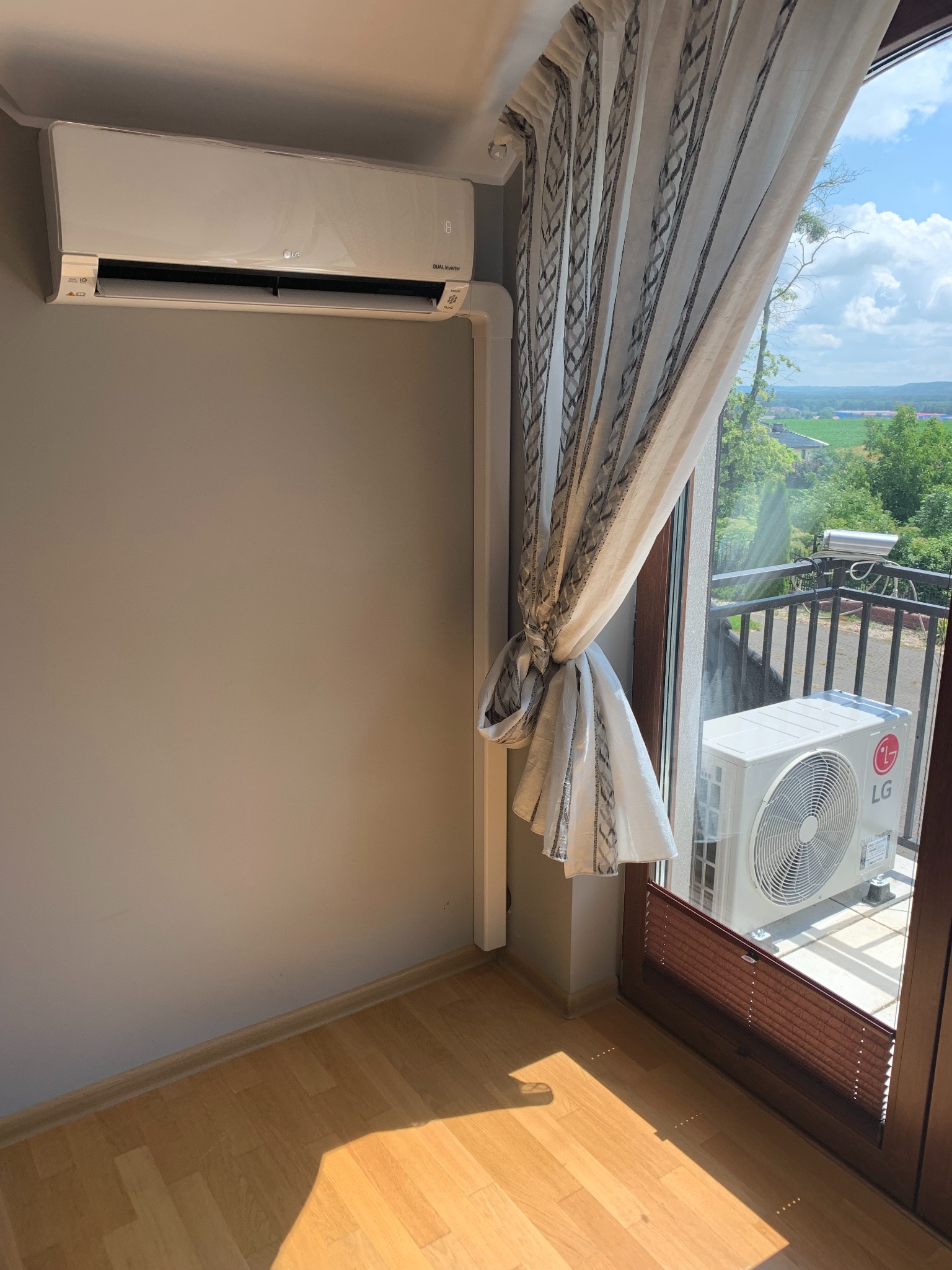 Zamontowana jednostka wewnętrzna klimatyzacji LG Dual Inverter na szarej ścianie, z widoczną jednostką zewnętrzną na balkonie za oknem z zasłoną.