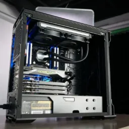 Otwarta obudowa komputera stacjonarnego z widocznym chłodzeniem wodnym, kartą graficzną GeForce GTX, pamięcią RAM z podświetleniem LED i zasilaczem Phanteks, ustawiona na stole z czarnym tłem.