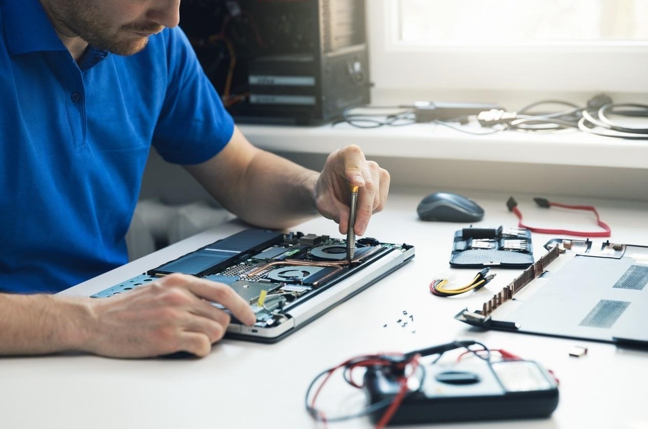 Serwisant w niebieskiej koszulce naprawia laptopa na białym stole w pobliżu okna, używając śrubokręta precyzyjnego; widoczny otwarty komputer, narzędzia i części.