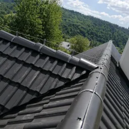 Czarny dach z dachówki ceramicznej z zamontowaną instalacją odgromową, widok z góry na tle zalesionych wzgórz i błękitnego nieba z obłokami.