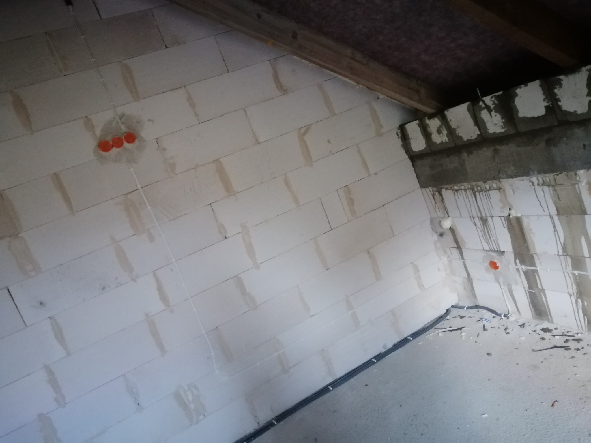 Surowa ściana z betonu komórkowego w trakcie prac instalacyjnych, widoczne przewody elektryczne prowadzone po ścianie i pomarańczowe puszki pod gniazdka.