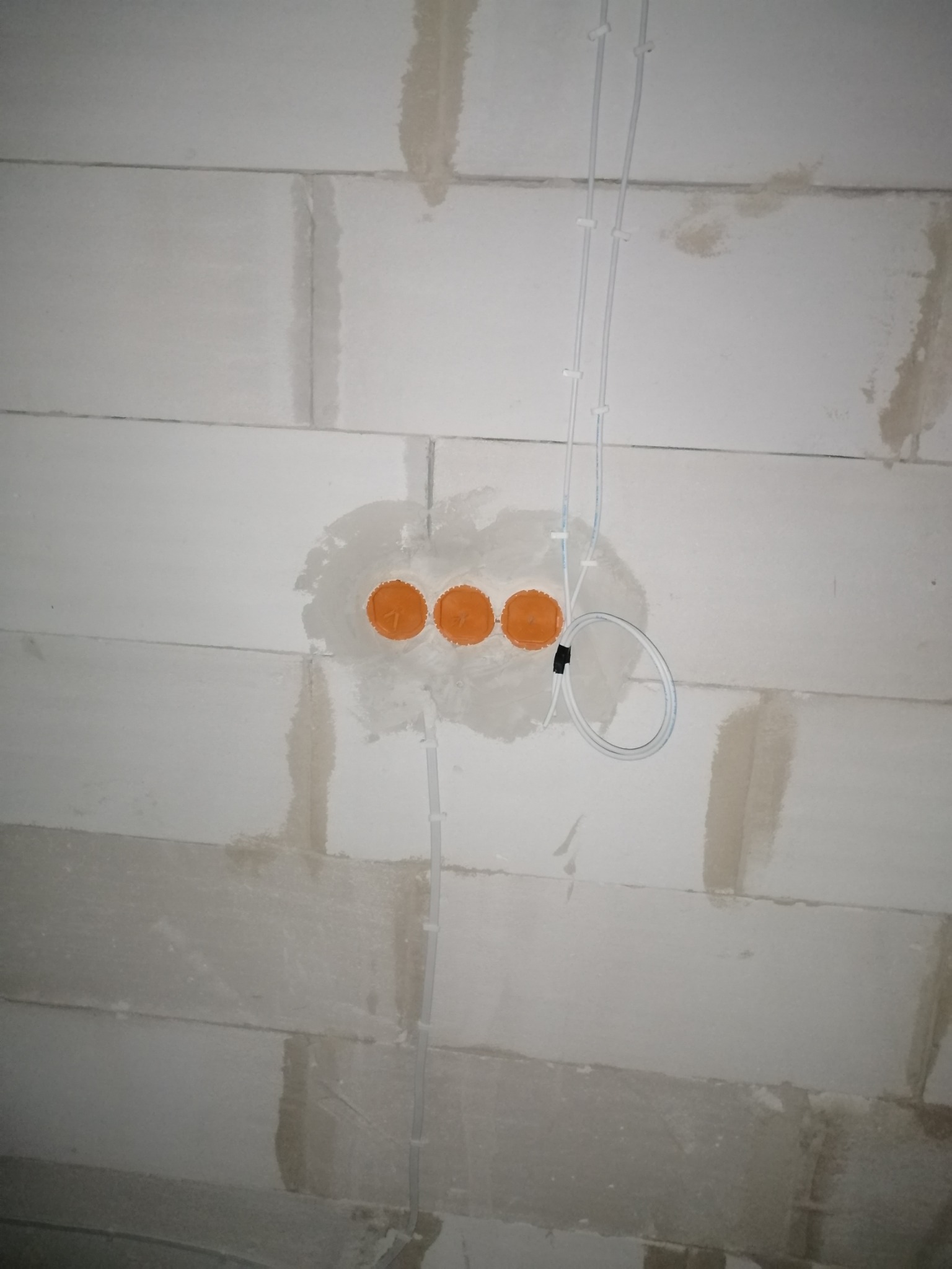 Ściana z betonu komórkowego z trzema pomarańczowymi puszkami podtynkowymi i przewodami elektrycznymi zamocowanymi do ściany za pomocą białych uchwytów.