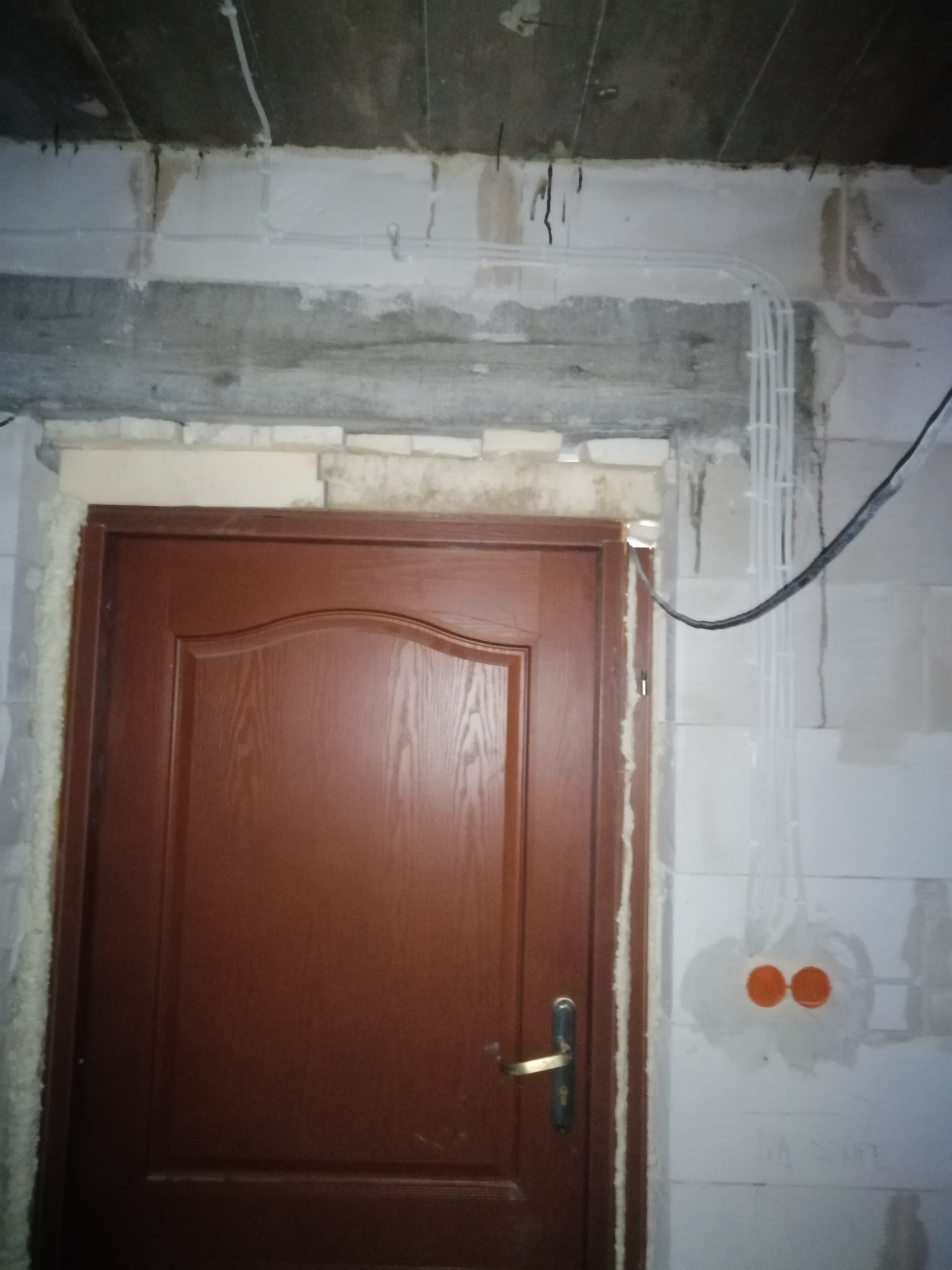 Wnętrze w trakcie remontu: widoczne białe przewody elektryczne w plastikowych rurkach mocowane na ścianie z białych bloczków, obok zamontowane brązowe drzwi z ościeżnicą uszczelnioną pianką...