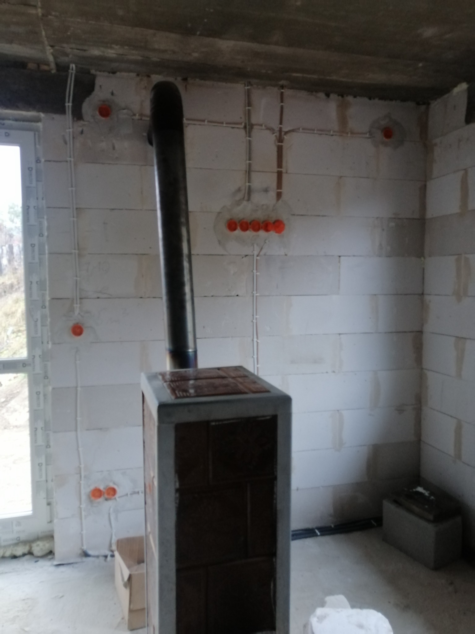 Surowy stan wnętrza z widoczną instalacją elektryczną w ścianach z betonu komórkowego, pomarańczowe puszki elektryczne oraz pionowy przewód kominowy pieca.