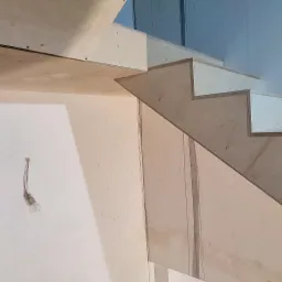 Surowe drewniane schody z płyty OSB w trakcie montażu, widoczna surowa ściana z wystającą żarówką, etap wykończeniowy.