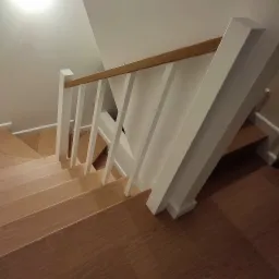 Widok z góry na drewniane schody z białymi balustradami i drewnianą poręczą, oświetlone ciepłym światłem.