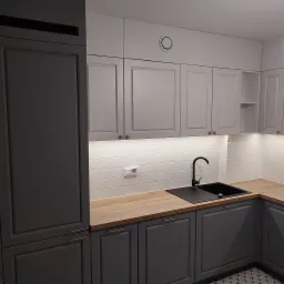 Nowoczesna kuchnia z szarymi szafkami z frezowanymi frontami, drewnianym blatem i czarnym zlewozmywakiem, oświetlona listwą LED pod szafkami górnymi.