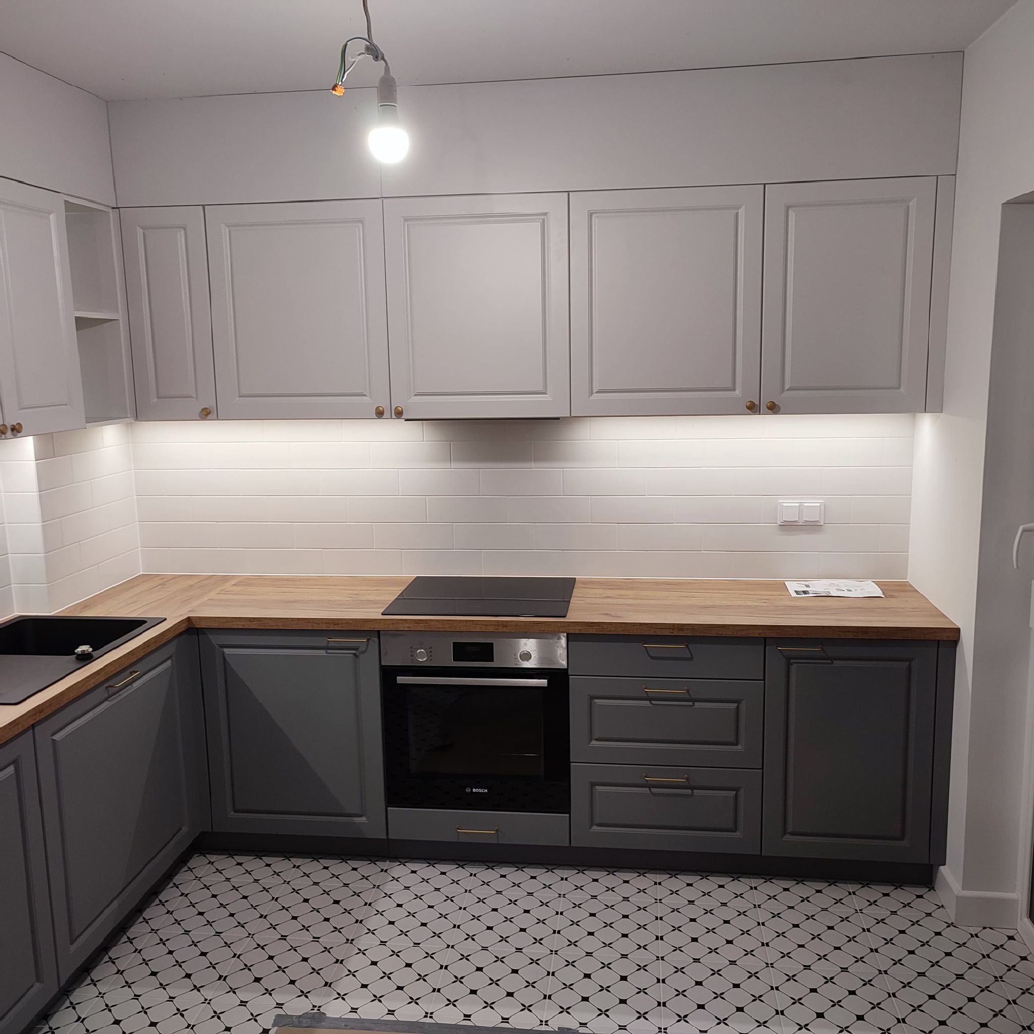 Szare szafki kuchenne z drewnianym blatem, białymi płytkami na ścianie i wzorzystą podłogą, widoczna płyta indukcyjna i piekarnik, niedokończone podłączenie oświetlenia sufitowego.
