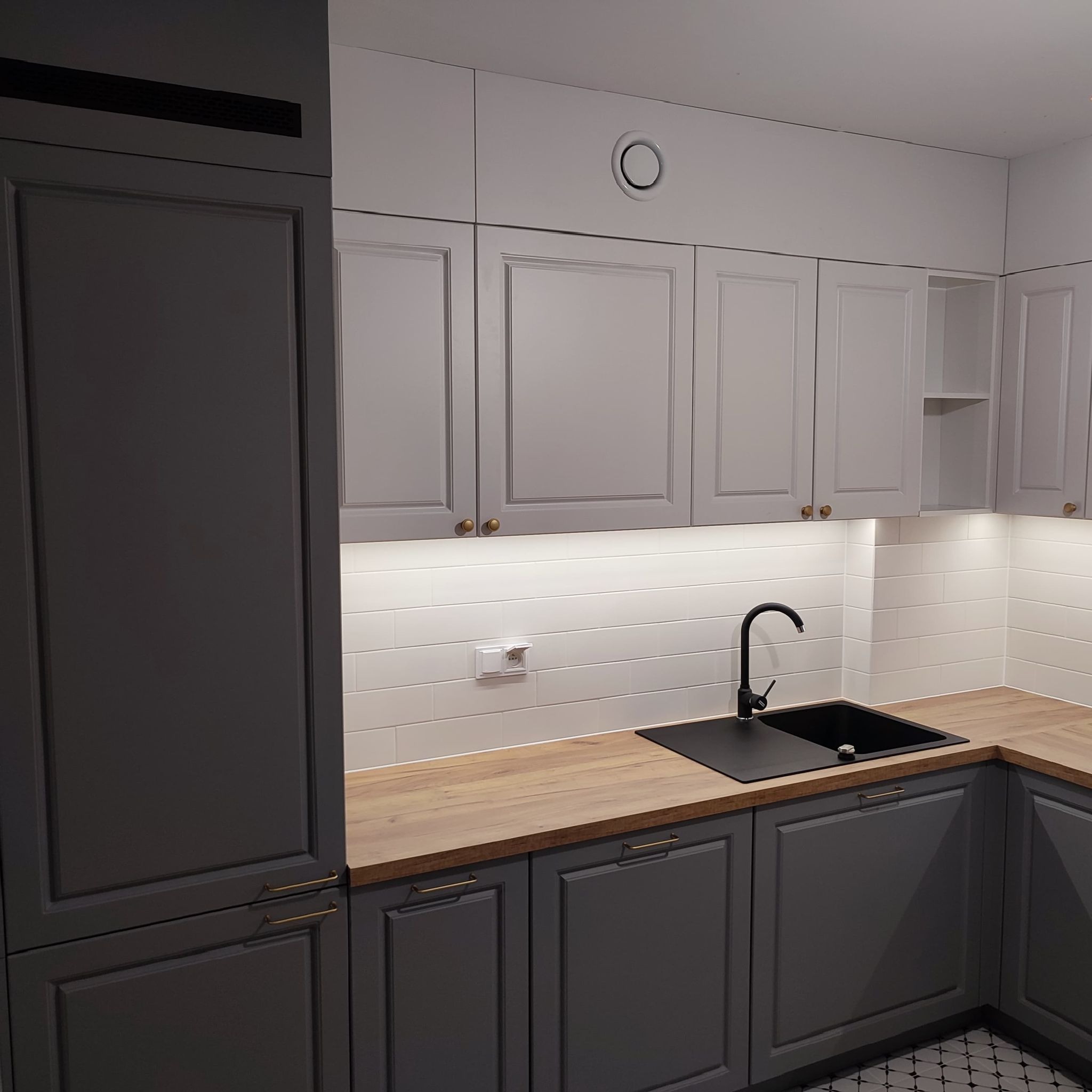 Nowoczesna kuchnia z szarymi szafkami z frezowanymi frontami, drewnianym blatem i czarnym zlewozmywakiem, oświetlona listwą LED pod szafkami górnymi.