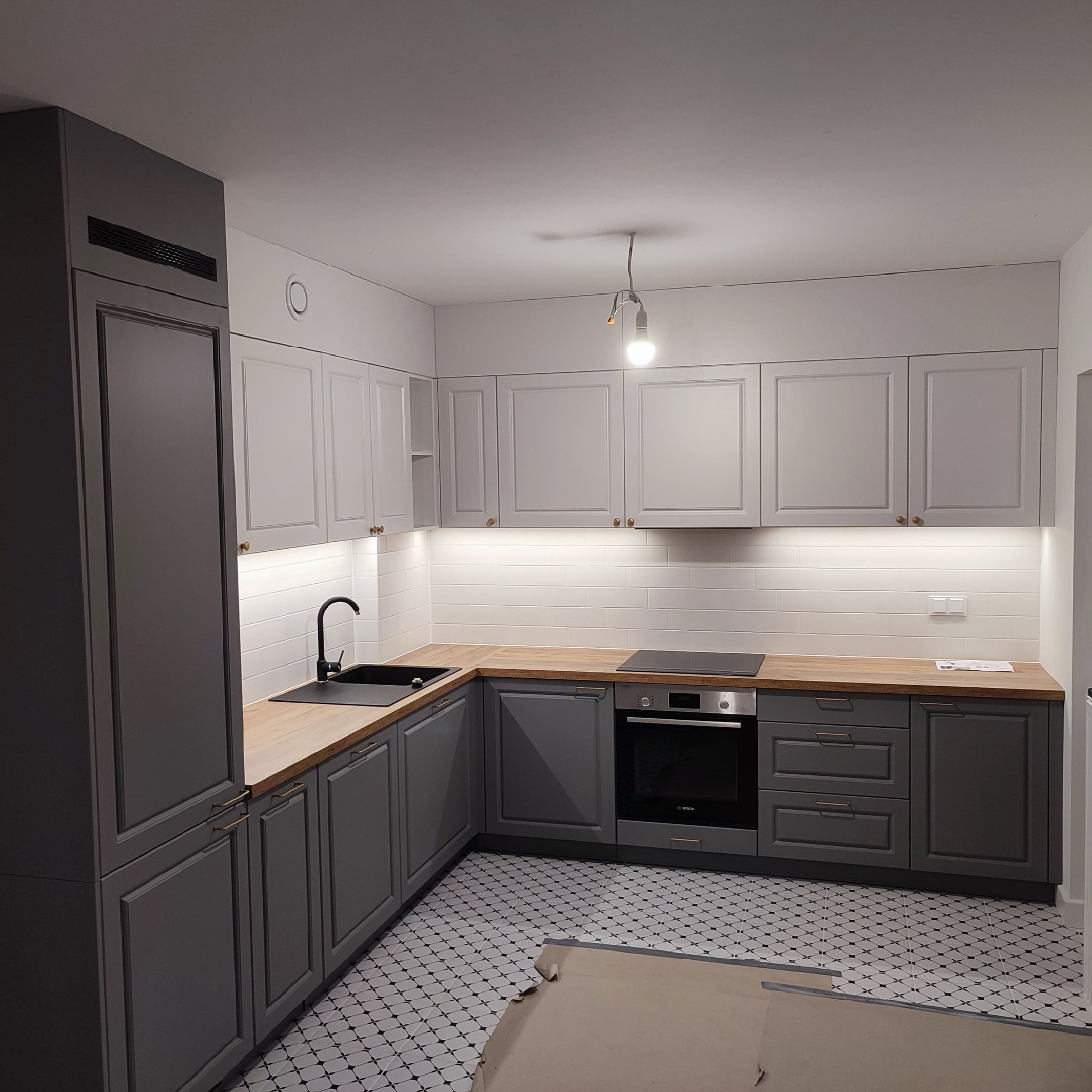 Nowoczesna, szara kuchnia z drewnianym blatem, czarnym zlewem i białą glazurą, oświetlona taśmą LED pod szafkami wiszącymi i z wzorzystą podłogą.