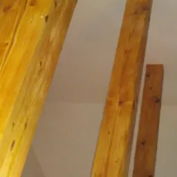 Poddasze w trakcie adaptacji: widoczne drewniane belki konstrukcyjne o ciepłym odcieniu, surowa ściana z płyt gipsowo-kartonowych przed szpachlowaniem, okno w tle.