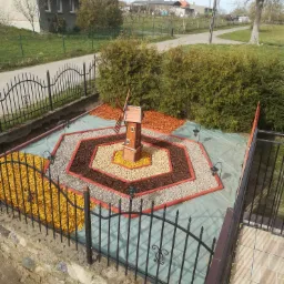 Ogródek przed domem z dekoracyjnym drewnianym wiatrakiem, otoczony geometrycznymi wzorami z kory, kamieni i żwirku, oświetlony małymi lampkami ogrodowymi, ogrodzony metalowym płotem.