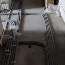 Szare rury instalacyjne ułożone równolegle na podłodze w surowym stanie, przymocowane do siebie białymi opaskami, z kartonem Tubolit w tle.