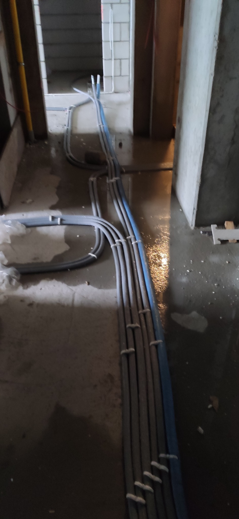 Szare i niebieskie rury instalacyjne prowadzone po mokrej posadzce w surowym wnętrzu budynku w trakcie budowy.