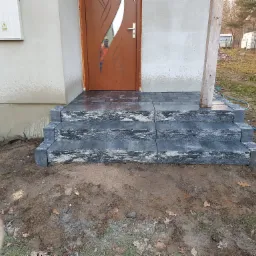 Schody wejściowe do domu w Poraju, wyłożone ciemną kostką granitową o nieregularnej fakturze, prowadzące do brązowych drzwi z przeszkleniem.