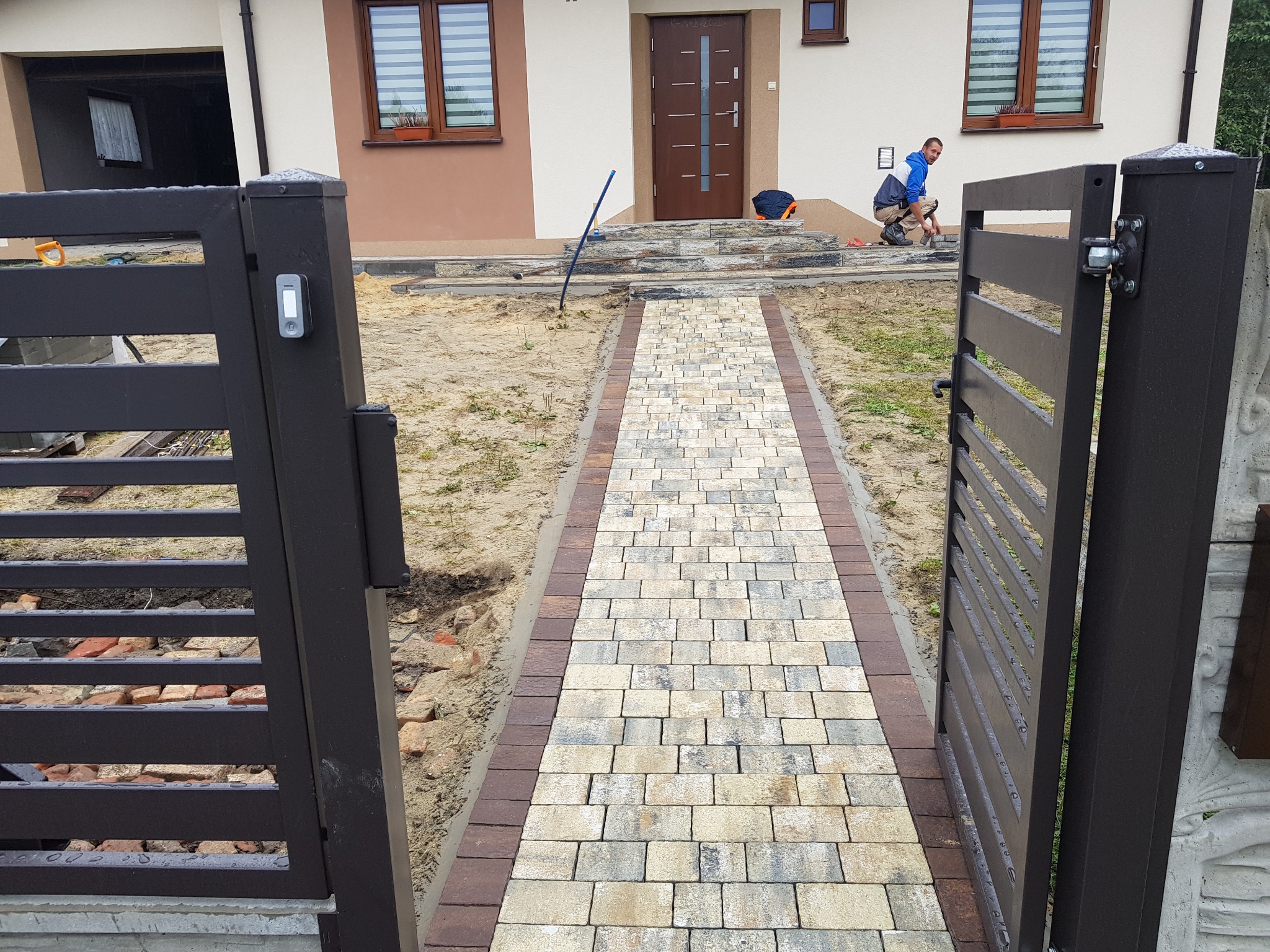 Nowo ułożony chodnik z kostki brukowej w odcieniach szarości i brązu prowadzący od furtki do wejścia domu, widoczny pracownik montujący obrzeża schodów wejściowych.