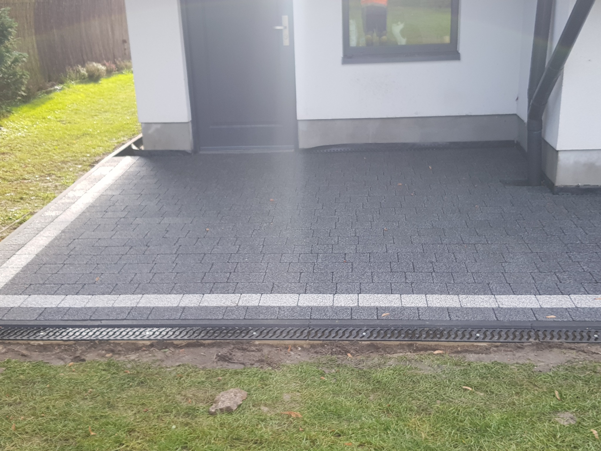 Ułożona szara kostka brukowa przed budynkiem z białymi ścianami, z odpływem liniowym i obrzeżem z jasnej kostki.