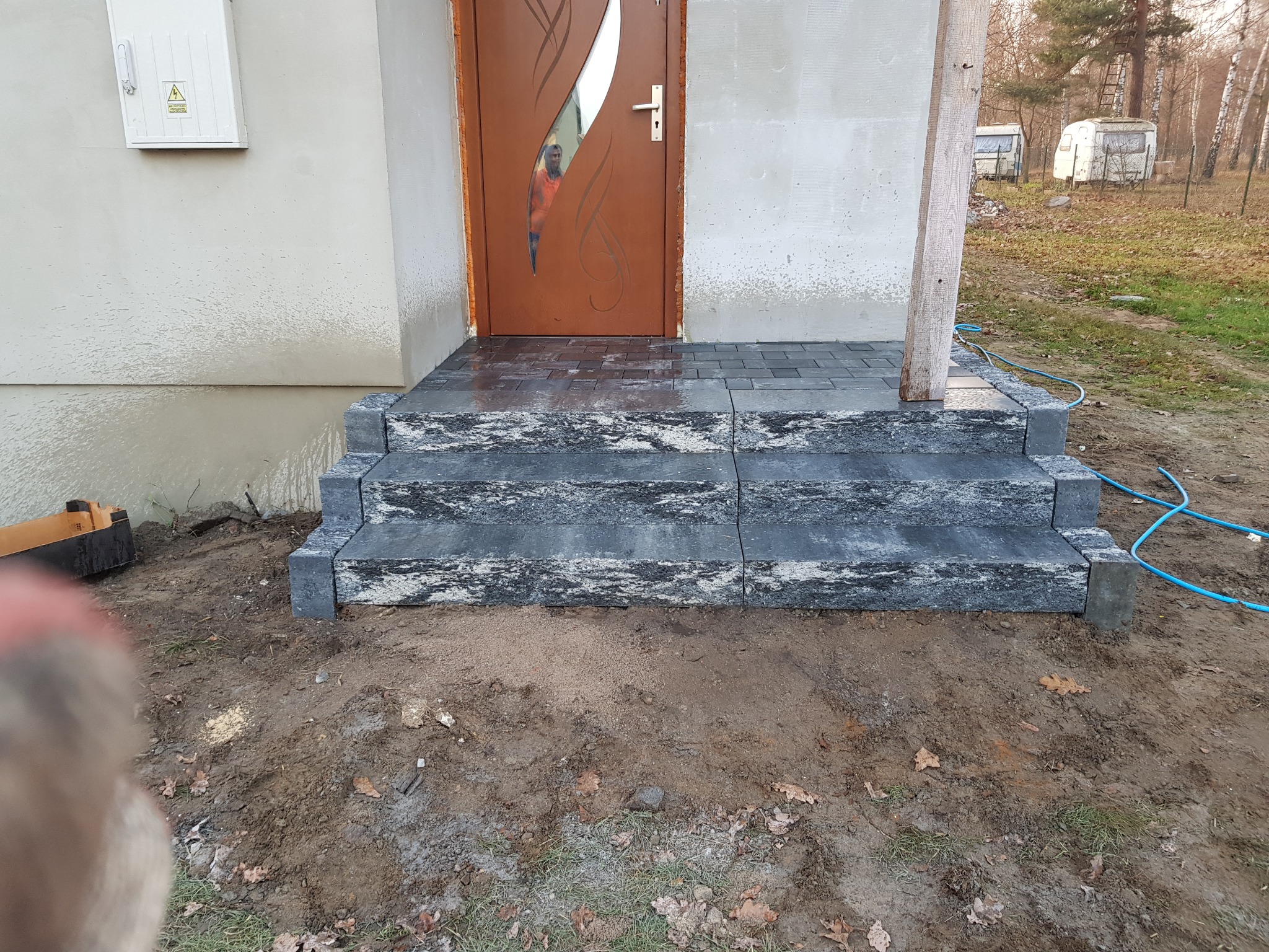 Schody wejściowe do domu w Poraju, wyłożone ciemną kostką granitową o nieregularnej fakturze, prowadzące do brązowych drzwi z przeszkleniem.