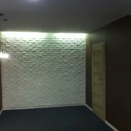 Wnętrze pokoju z dekoracyjną ścianą z białej cegły, oświetloną od góry, ciemna ściana boczna i drzwi.