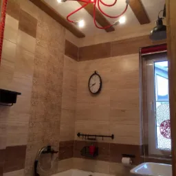 Wykończona łazienka z beżowymi płytkami, białą wanną z czerwonym akcentem, umywalką, dekoracyjnym oświetleniem sufitowym z czerwonymi kablami i drewnianymi belkami, uzupełniona o czarny zegar ścienny.